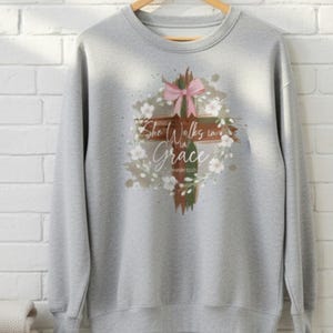 May include: A pale grey sweatshirt featuring a floral cross design. The cross is decorated with a pink bow, white flowers, and the text "She Walks in Grace". The sweatshirt is hanging on a wooden hanger.