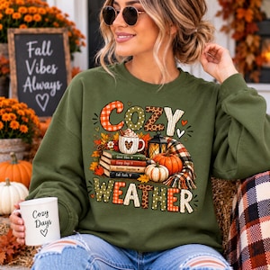 May include: An olive green long-sleeve sweatshirt featuring a graphic print with the text "COZY WEATHER". The design includes a stack of books, a mug with a heart, a lantern, pumpkins, and a plaid scarf. The sweatshirt has a crew neckline.