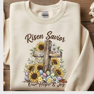 Puede incluir: Sudadera color crema con una cruz de madera adornada con girasoles, margaritas y huevos de Pascua. Los textos "Risen Savior" y "Our Hope & Joy" están impresos encima y debajo de la cruz. La sudadera está doblada sobre una superficie de madera.
