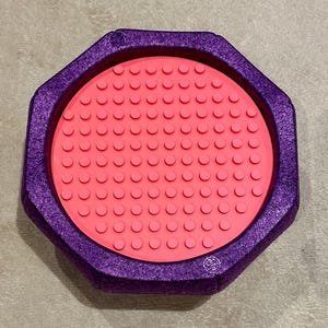 Op de afbeelding: Een paarse achthoekige tray met een getextureerd oppervlak omringt een cirkelvormige, koraalkleurige basis. De basis heeft talrijke kleine, verhoogde cirkels. De tray is ontworpen om kleine items in te bewaren.