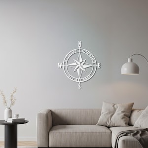 Op de afbeelding: Een wit metalen kompasroos wandkunstwerk. Het kompas heeft een ster in het midden, met richtingaanwijzers en tekst rondom de omtrek. Het stuk is bevestigd aan een grijze muur, boven een bank.