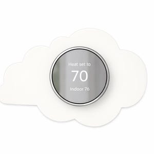 May include: A smart thermostat with a silver dial, set to 70 degrees, is mounted on a white cloud-shaped backing. The text on the dial reads "Heat set to 70 Indoor 76". The design is modern and minimalist.