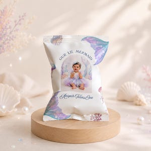 May include: A custom snack bag featuring a white background with purple and blue mermaid tail illustrations, coral, and shells. The centre displays a photograph of a baby in a tutu with the text "OUR LIL MERMAID" and "Aniyah Turns One" in blue.
