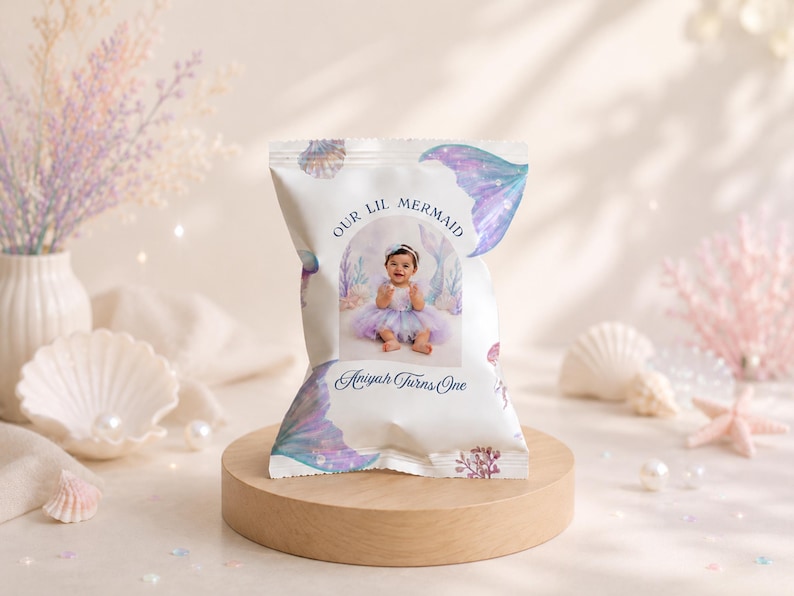 May include: A custom snack bag featuring a white background with purple and blue mermaid tail illustrations, coral, and shells. The centre displays a photograph of a baby in a tutu with the text "OUR LIL MERMAID" and "Aniyah Turns One" in blue.