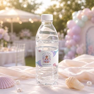 May include: A clear plastic water bottle with a white cap and a custom label featuring a photograph of a child in a tutu, the text "OUR LIL MERMAID" and "Alayna turns One", decorated with illustrations of colourful mermaid tails and seashells.