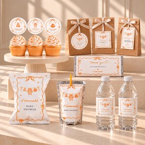 May include: A collection of baby shower party favours featuring orange and white themed decorations. Items include three cupcakes with circular toppers, three brown paper gift bags, a candy bar, a snack bag, a drink pouch, and two water bottles.
