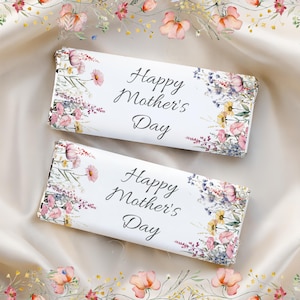 Puede incluir: Dos envoltorios blancos de barras de chocolate con la inscripción "Happy Mother's Day". Diseños florales en rosa, morado y amarillo adornan los bordes. Las barras descansan sobre una tela color crema, rodeadas por un borde floral.