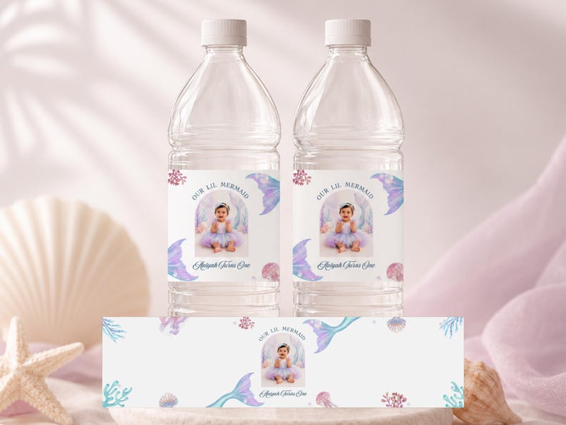 May include: Two clear plastic water bottles and a flat label featuring a photo of a baby in a tutu, surrounded by watercolour illustrations of mermaid tails, coral, and seashells. The text reads Our Lil Mermaid and Aaliyah Turns One.