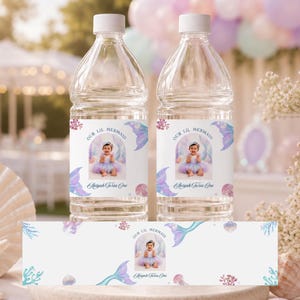 May include: Two clear plastic water bottles with custom labels featuring a photo of a child, the text Our Lil Mermaid and Aaliyah Turns One, decorated with watercolour illustrations of mermaid tails, seashells, and coral in shades of purple and blue.