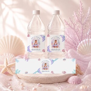 May include: Two clear plastic water bottles with white caps and custom labels featuring a photograph of a child in a mermaid costume, the text Our Lil Mermaid, and the name Aaliyah Turns One, alongside a matching horizontal label strip.