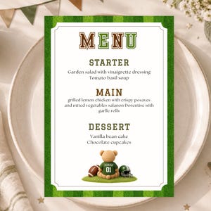 May include: A rectangular menu card with a green grass-patterned border. The text reads MENU, STARTER, Garden salad with vinaigrette dressing, Tomato basil soup, MAIN, grilled lemon chicken with crispy posaves and mtted vegetables salanon Dorentise with garllc rolls, DESSERT, Vanilla bean cake, Chocolate cupcakes, and an illustration of a teddy bear wearing a green jersey with the name JONAH and number 01, flanked by a football and a helmet.