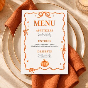 May include: A rectangular white menu card featuring an orange border with ribbon bows, small hearts, and star accents. The text reads MENU, APPETIZERS, Fresh Garden Salad, Tomato Basil Soup, ENTRÉES, Grilled Lemon Herb Chicken, Baked Salmon with Seasonal Vegetables, DESSERTS, Vanilla bean cake, Chocolate cupcakes, and includes a small orange pumpkin illustration.