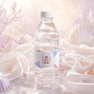 May include: A clear plastic water bottle with a white cap and a custom label featuring a photograph of a child in a mermaid costume, the text "OUR LIL MERMAID" and "Abigail Faith Eve", decorated with illustrations of mermaid tails and seashells.