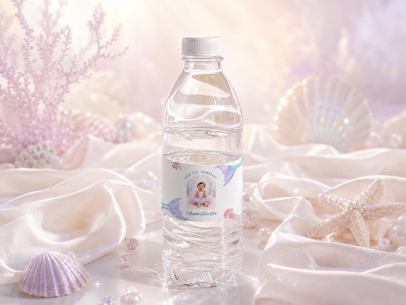 May include: A clear plastic water bottle with a white cap and a custom label featuring a photograph of a child in a mermaid costume, the text "OUR LIL MERMAID" and "Abigail Faith Eve", decorated with illustrations of mermaid tails and seashells.