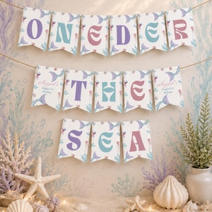 K&ouml;nnte beinhalten: Ein dekoratives Banner-Set mit Wimpeln in einem Meerjungfrauen-Muster und pastellfarbenen Buchstaben, die ONEDER THE SEA bilden. Die ersten und letzten Wimpel in der zweiten Reihe enthalten den Text Aniyah's Magical 1st Birthday in Schreibschrift.