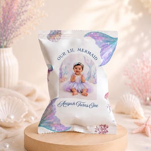 May include: A white snack bag featuring a watercolour mermaid theme with purple and blue tail illustrations, seashells, and coral. The centre displays a photograph of a baby in a tutu with the text "OUR LIL MERMAID" and "Aniyah Turns One".