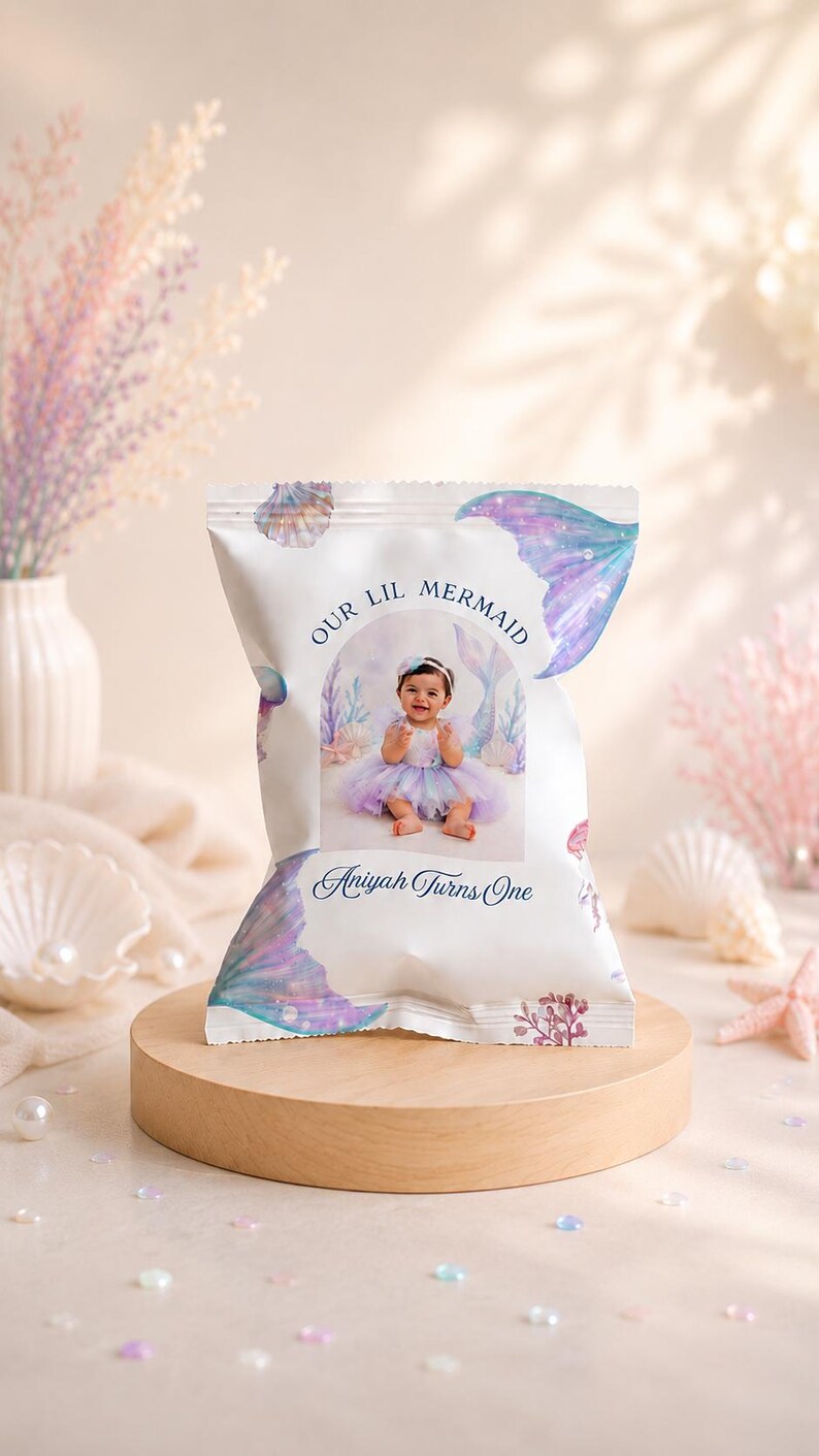 May include: A white snack bag featuring a watercolour mermaid theme with purple and blue tail illustrations, seashells, and coral. The centre displays a photograph of a baby in a tutu with the text "OUR LIL MERMAID" and "Aniyah Turns One".