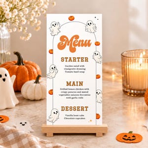 May include: A vertical rectangular menu card standing on a light wood base. The white card features orange text reading Menu, Starter, Main, and Dessert, with black text listing food items. It is decorated with small ghost and pumpkin illustrations.
