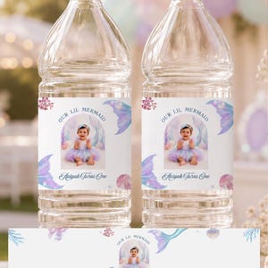 May include: Two clear plastic water bottles with custom labels featuring a photo of a baby in a mermaid outfit, the text "OUR LIL MERMAID" and "Aaliyah Turns One," surrounded by watercolour illustrations of mermaid tails, coral, and seashells.