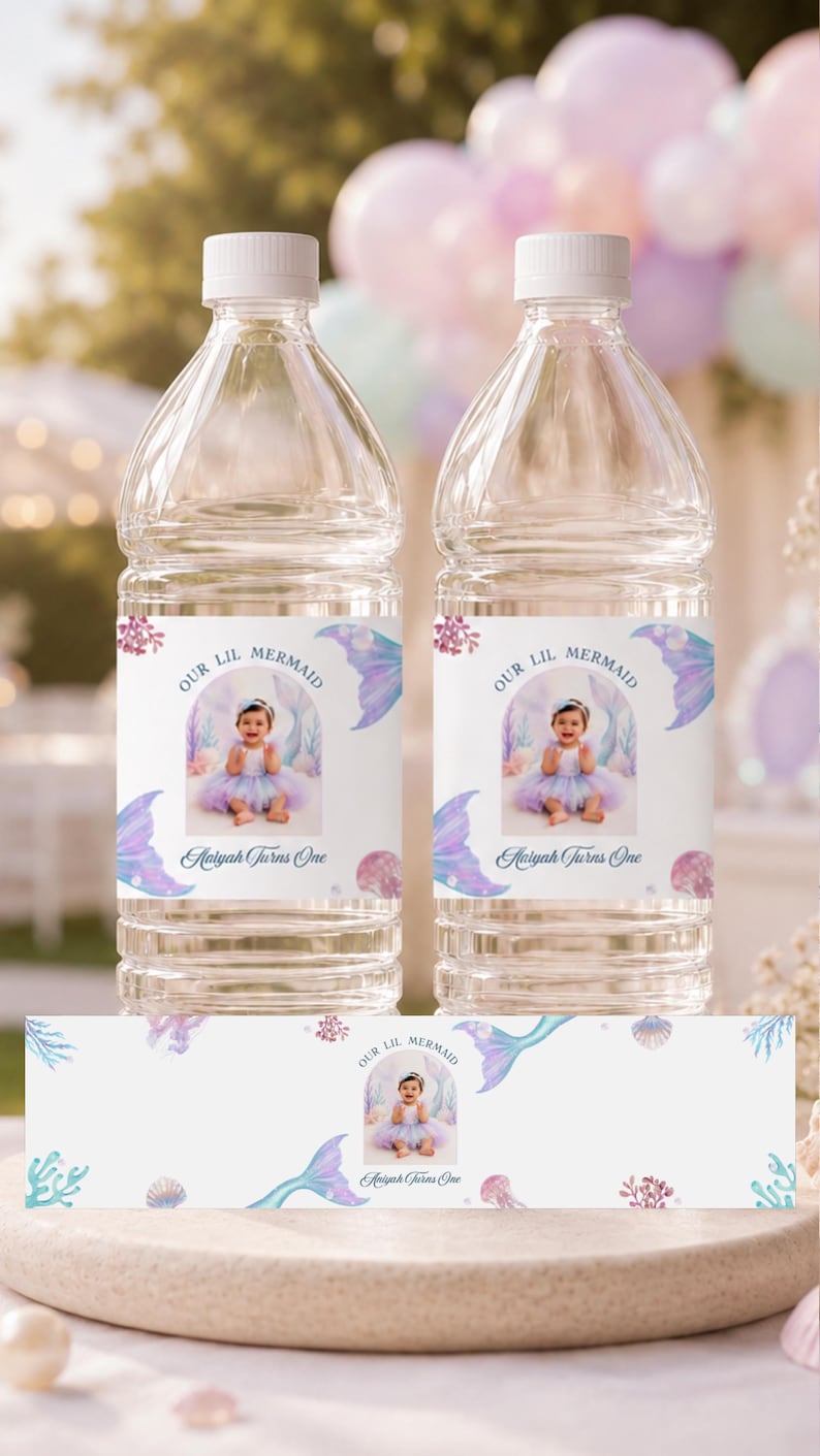 May include: Two clear plastic water bottles with custom labels featuring a photo of a baby in a mermaid outfit, the text "OUR LIL MERMAID" and "Aaliyah Turns One," surrounded by watercolour illustrations of mermaid tails, coral, and seashells.