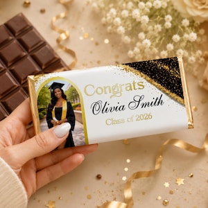 May include: A chocolate bar with a personalized wrapper. The wrapper features a photo of a graduate in a cap and gown, with the text "Congrats Olivia Smith Class of 2026" in gold lettering. A dark chocolate bar is visible in the background.