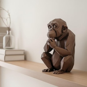 Könnte beinhalten: Eine braune, geometrische Schimpansen-Skulptur sitzt auf einem Holzregal. Die Skulptur befindet sich in sitzender Position mit unter dem Kinn gefalteten Händen. Eine Vase mit Zweig und Bücher befinden sich auf dem Regal.