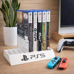 Könnte beinhalten: Ein weißer PS5-Spielhalter mit dem PS5-Logo und einer Reihe von Spielhüllen. Die Spiele umfassen "Hogwarts Legacy", "FC24" und "Gran Turismo 7". Zwei Gamecontroller, einer blau und einer orange, befinden sich im Vordergrund.