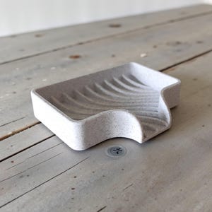 May include: A speckled, light gray soap dish with a curved design. The dish has a rectangular shape with a wavy, sloped edge for drainage. The interior features curved ridges. The soap dish is made of a solid material.