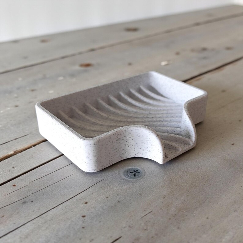 May include: A speckled, light gray soap dish with a curved design. The dish has a rectangular shape with a wavy, sloped edge for drainage. The interior features curved ridges. The soap dish is made of a solid material.
