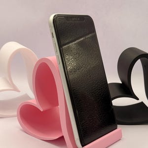 Könnte beinhalten: Ein rosa herzförmiger Telefonständer, der ein schwarzes Smartphone hält. Im Hintergrund sind ein weißer und ein schwarzer herzförmiger Ständer zu sehen. Der Telefonständer besteht aus einem glatten, festen Material.
