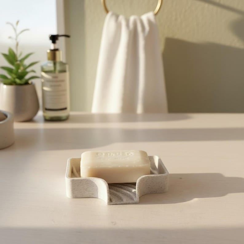 May include: A light-colored soap bar rests in a speckled, light gray soap dish. The dish has a unique, curved design. A bottle of liquid soap and a small potted plant are in the background. A white towel hangs on a gold ring.