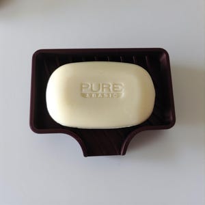 May include: A bar of soap with the words "PURE & BASIC" embossed on it, resting in a dark brown soap dish. The soap is a light cream color and has a smooth, rounded shape. The soap dish has a ridged design.