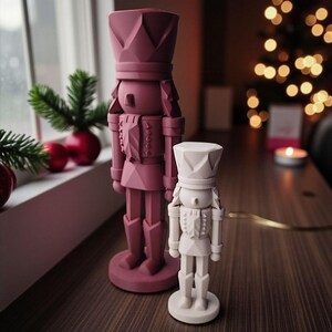 May include: Two geometric nutcracker figurines standing upright. The larger nutcracker is a deep mauve colour, while the smaller nutcracker is a solid off-white. Both figures feature faceted, angular surfaces and are mounted on circular bases.