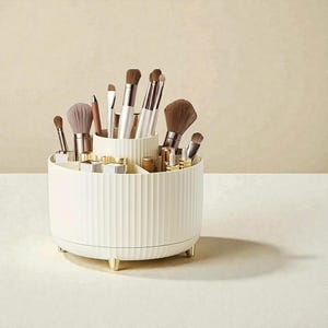 Op de afbeelding: Een cr&egrave;mekleurige, roterende make-up organizer met verticale geribbelde details. Het heeft meerdere compartimenten en bevat diverse make-up borstels, lippenstiften en andere schoonheidsproducten. De organizer heeft goudkleurige voetjes en accenten.