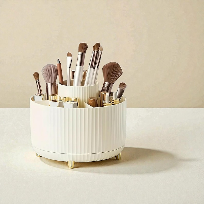 Op de afbeelding: Een cr&egrave;mekleurige, roterende make-up organizer met verticale geribbelde details. Het heeft meerdere compartimenten en bevat diverse make-up borstels, lippenstiften en andere schoonheidsproducten. De organizer heeft goudkleurige voetjes en accenten.