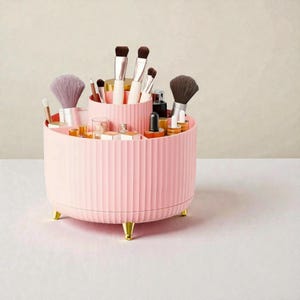 Op de afbeelding: Een roze, geribbelde, roterende make-up organizer met goudkleurige voetjes. De organizer is gevuld met diverse make-up kwasten, flesjes en containers. De organizer heeft twee trapsgewijze secties.