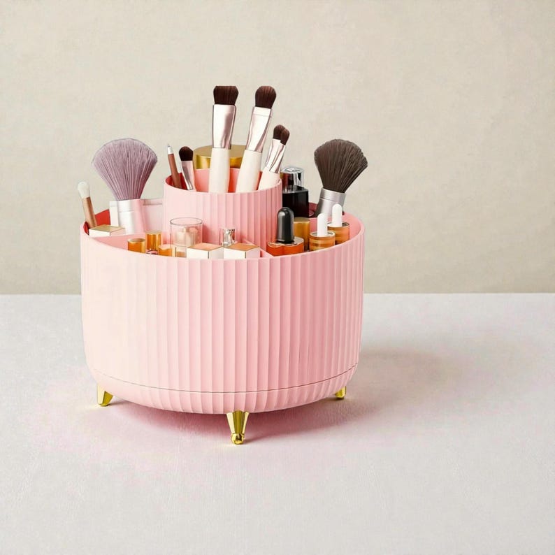 Op de afbeelding: Een roze, geribbelde, roterende make-up organizer met goudkleurige voetjes. De organizer is gevuld met diverse make-up kwasten, flesjes en containers. De organizer heeft twee trapsgewijze secties.