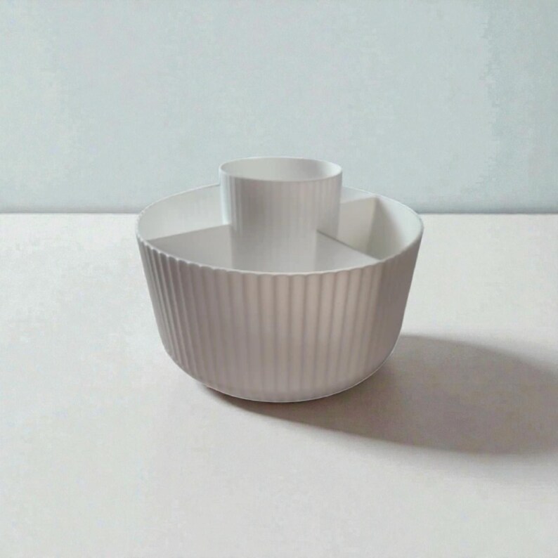 Op de afbeelding: Een witte, geribbelde keramische serveerschaal met een centraal cilindrisch compartiment. De kom heeft een verdeeld interieur en is ontworpen voor het serveren van snacks of hapjes. De kom staat op een wit oppervlak tegen een lichtblauwe achtergrond.