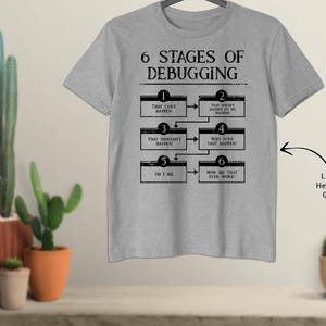 May include: Light heather grey t-shirt featuring the text "6 STAGES OF DEBUGGING" and a flowchart illustrating debugging steps. The shirt is displayed on a wooden surface with potted cacti in the background.