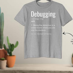 May include: Light heather grey t-shirt with the definition of "Debugging" printed in white. The definition includes the pronunciation, part of speech, and a humorous explanation. The shirt is displayed on a hanger.