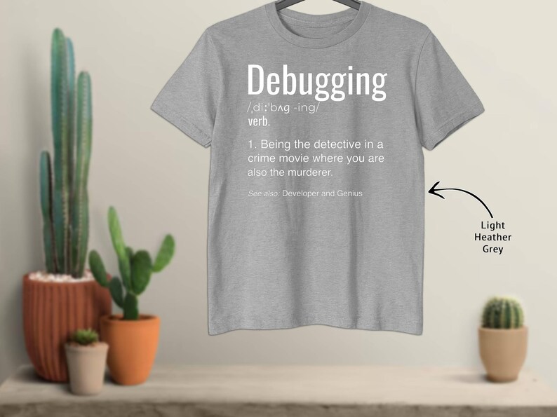 Może przedstawiać: Jasnoszara koszulka z nadrukiem definicji słowa "Debugging" w kolorze białym. Definicja zawiera wymowę, część mowy i humorystyczne wyjaśnienie. Koszulka jest prezentowana na wieszaku.
