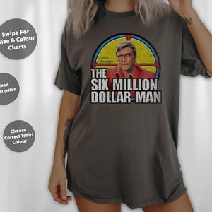 May include: Dark grey t-shirt with a graphic of "The Six Million Dollar Man", featuring a portrait of the character in a red shirt. The text "THE SIX MILLION DOLLAR MAN" is printed in white. The shirt has a vintage, worn appearance.