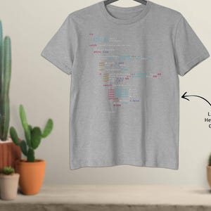 May include: Light heather grey t-shirt featuring a colourful code script design. The text is in blue, red, and white. The shirt is hanging on a hanger. The background includes cacti in terracotta pots.