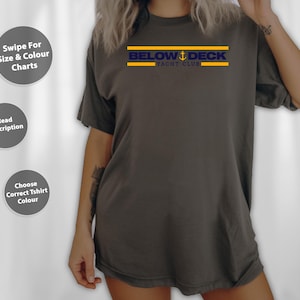 May include: A dark grey t-shirt featuring the text "BELOW DECK YACHT CLUB" in navy blue and gold. The shirt has a loose fit and short sleeves. The image also includes text prompts for size and colour charts and description.