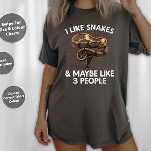 May include: Dark grey t-shirt featuring the text "I LIKE SNAKES & MAYBE LIKE 3 PEOPLE" and a graphic of a brown and tan snake. The shirt also has three grey circular buttons with text.