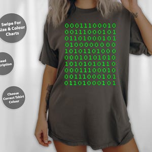 May include: Dark grey t-shirt with green binary code design. The shirt features a series of green binary code numbers. The shirt has a relaxed fit and short sleeves. The text on the shirt reads "00011100010".