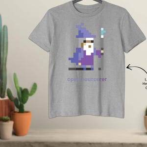 May include: Light heather grey t-shirt with a pixelated wizard design in purple and blue, holding a staff. The text "open sourcerer" is below the graphic. The shirt is displayed on a hanger.