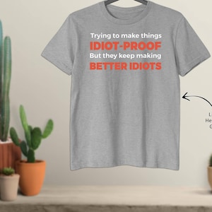 May include: Light heather grey t-shirt with the text "Trying to make things IDIOT-PROOF But they keep making BETTER IDIOTS" in white and red. The shirt is hanging on a hanger.