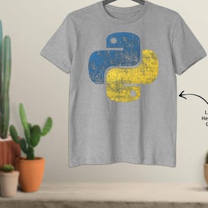 May include: Light heather grey t-shirt with a distressed blue and yellow Python logo. The shirt is hanging on a hanger. The background features potted cacti and succulents.