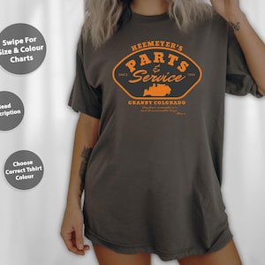 May include: Dark grey t-shirt featuring an orange graphic with the text "HEEMEYER'S PARTS & SERVICE GRANBY COLORADO." The design includes a bulldozer illustration. The shirt has a loose fit.